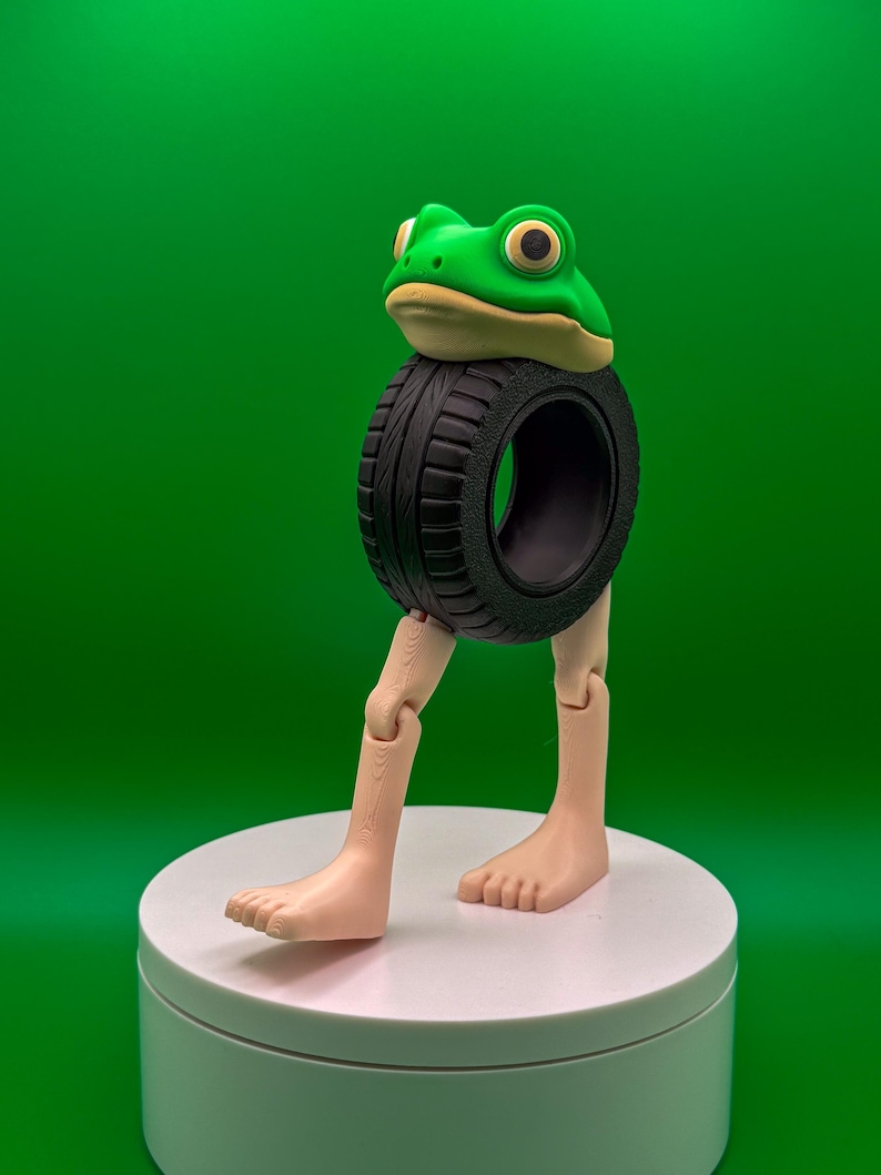 May include: A whimsical sculpture of a frog head atop a black tire, with articulated legs and feet. The frog is green with yellow eyes and a tan underside. The sculpture is on a white platform against a green backdrop.