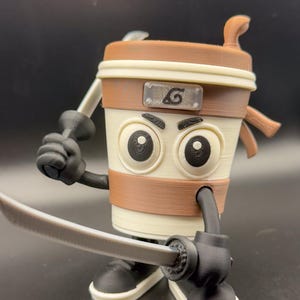 May include: A 3D-printed coffee cup character with a brown and beige color scheme, holding two toy swords. The cup has large, cartoonish eyes, a brown band, and a symbol on the front. The character is posed as if ready for battle.
