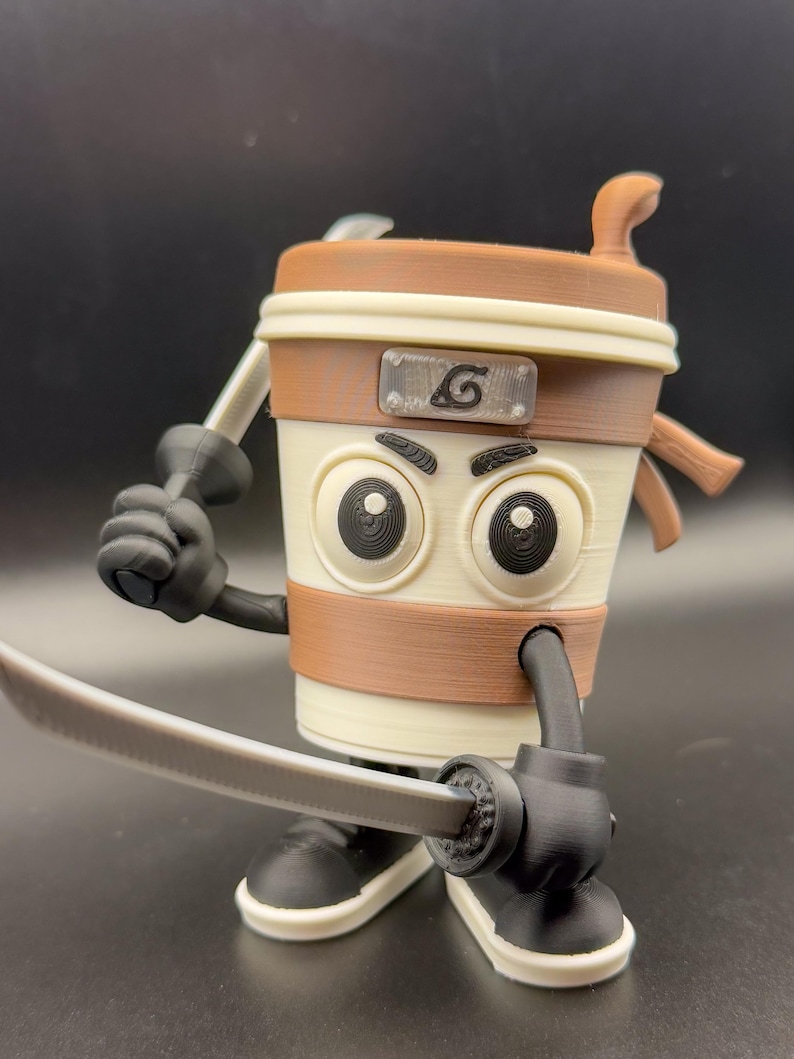 May include: A 3D-printed coffee cup character with a brown and beige color scheme, holding two toy swords. The cup has large, cartoonish eyes, a brown band, and a symbol on the front. The character is posed as if ready for battle.