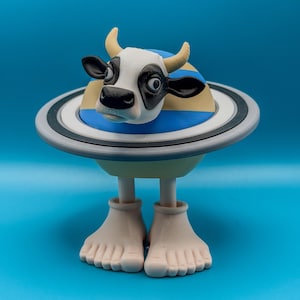 May include: A whimsical sculpture of a cow with a black and white face, yellow horns, and a blue and yellow striped body, set within a Saturn-like ring. The sculpture stands on two beige feet against a blue background.