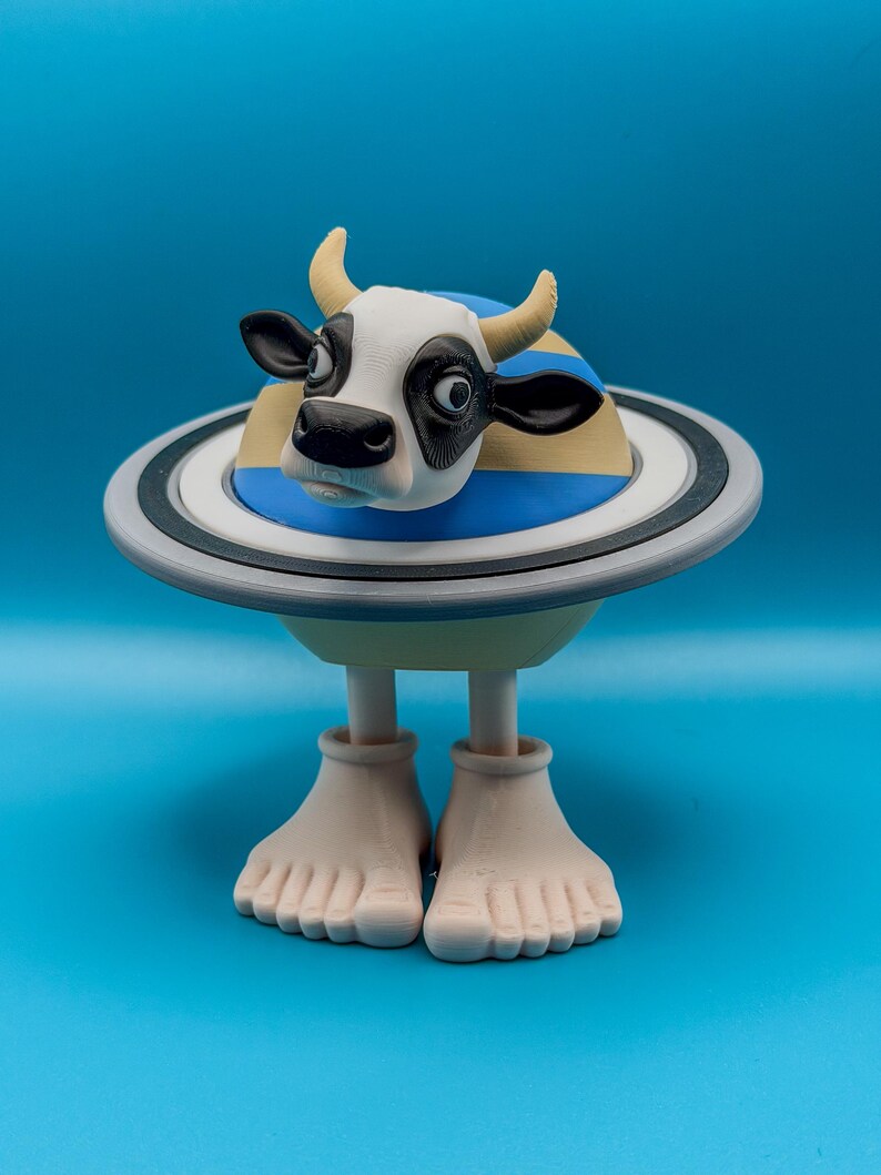 May include: A whimsical sculpture of a cow with a black and white face, yellow horns, and a blue and yellow striped body, set within a Saturn-like ring. The sculpture stands on two beige feet against a blue background.