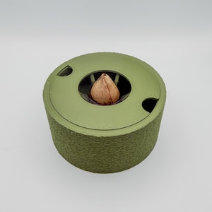 Può includere: Un contenitore cilindrico verde salvia con coperchio. Il coperchio ha un'apertura circolare che rivela un oggetto marrone. L'esterno del contenitore ha un aspetto strutturato e stratificato. L'oggetto è posto su uno sfondo bianco.
