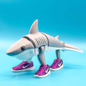 Pode incluir: Uma figura de tubarão impressa em 3D em tons de cinza e branco, usando tênis roxos com solas brancas e um logotipo branco. O tubarão tem uma barbatana, cauda e corpo articulado. O fundo é azul claro sólido.