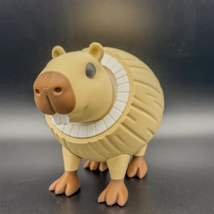 May include: A tan-colored capybara figurine with a detailed textured body, brown nose, and white teeth. The figurine has a white collar and small, dark eyes. The capybara is standing on four brown feet.