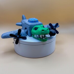 May include: A whimsical toy airplane featuring a green alligator head as the plane's nose. The gray plane has black propellers and a blue cockpit. The toy is displayed on a white, round platform against a neutral background.