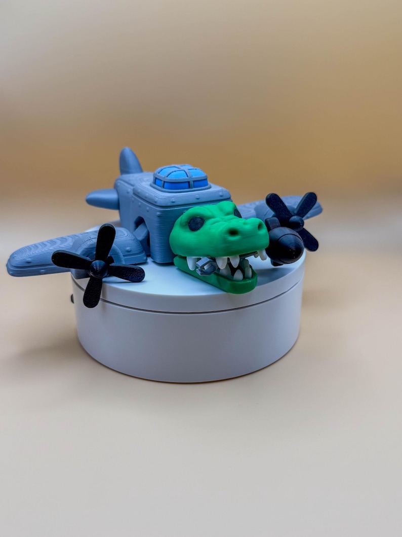 May include: A whimsical toy airplane featuring a green alligator head as the plane's nose. The gray plane has black propellers and a blue cockpit. The toy is displayed on a white, round platform against a neutral background.