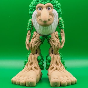May include: A whimsical, 3D-printed figure with a tree-like design. The figure has a green, leafy head and arms, a white beard, and a long nose. The body and feet are tan, resembling tree bark. The figure stands on a green background.