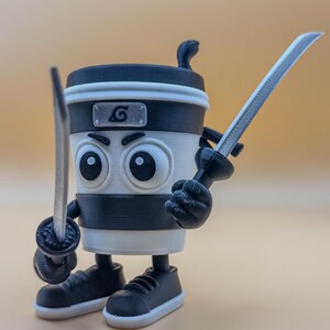 May include: A 3D-printed coffee cup character with large eyes, a black and white color scheme, and a ninja headband. The cup is holding two toy swords and wearing black sneakers. A silver emblem with a swirl is on the cup.