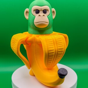 May include: A 3D-printed sculpture of a monkey head emerging from a yellow banana peel. The monkey's head is green with a tan face and ears. The banana peel is bright yellow with a black tip. The sculpture is on a white base against a green background.
