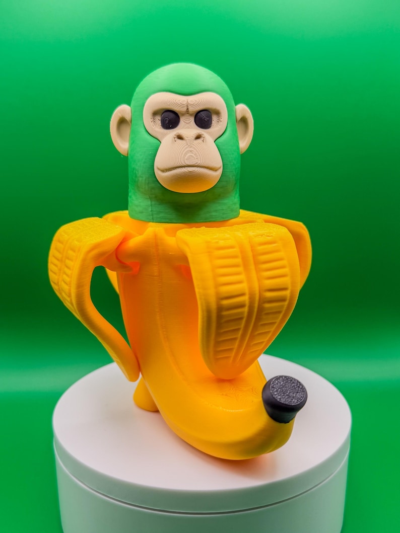 May include: A 3D-printed sculpture of a monkey head emerging from a yellow banana peel. The monkey's head is green with a tan face and ears. The banana peel is bright yellow with a black tip. The sculpture is on a white base against a green background.