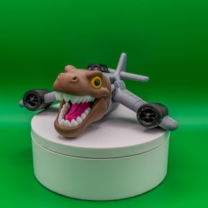 May include: A toy dinosaur head with a wide open mouth, white teeth, and a pink tongue, attached to a gray airplane body. The toy has black wheels and is set against a green background.
