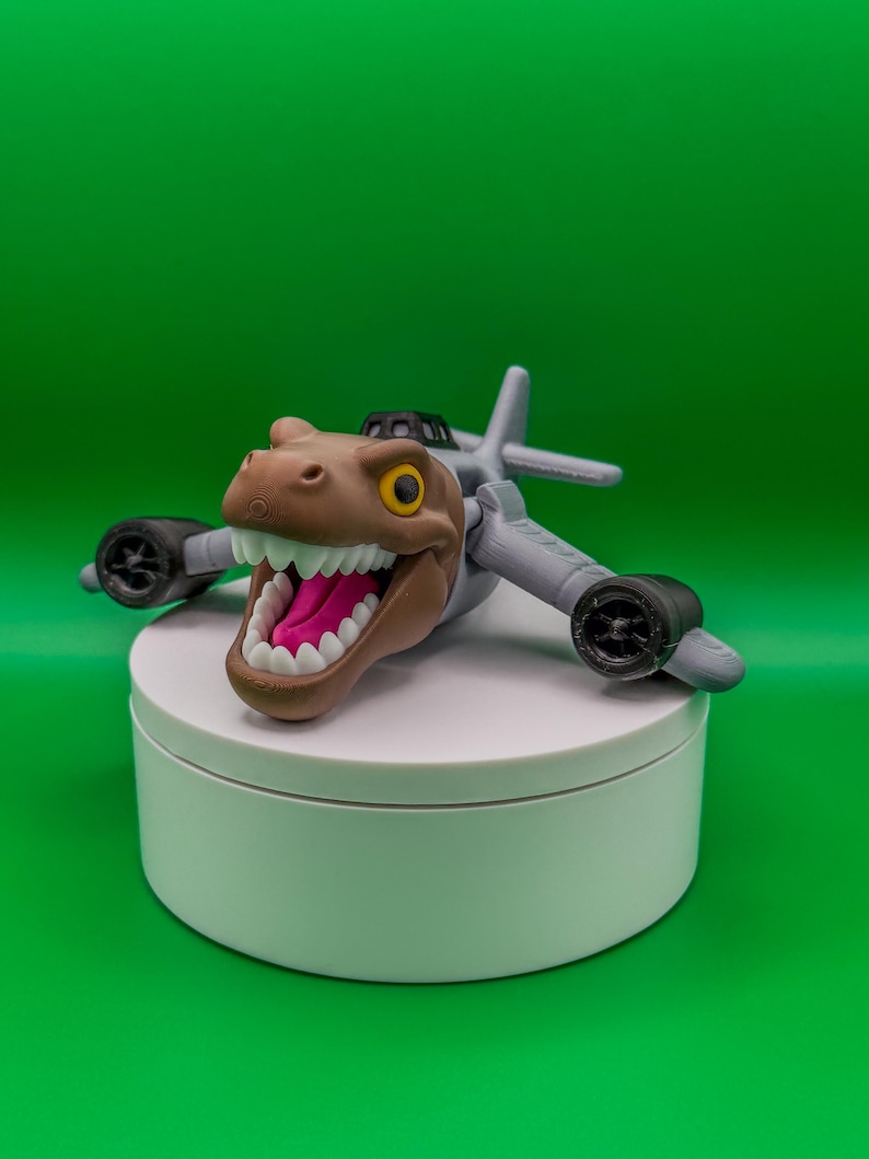 May include: A toy dinosaur head with a wide open mouth, white teeth, and a pink tongue, attached to a gray airplane body. The toy has black wheels and is set against a green background.