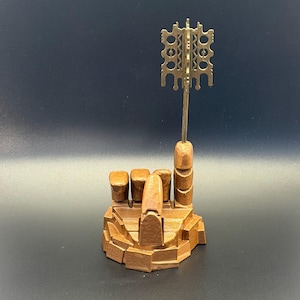 May include: A brown, hand-shaped sculpture with fingers curled into a fist, except for the index finger which is extended upwards. A metallic, gold-coloured, flat, rectangular key-like object with intricate cut-out patterns is attached to the finger.