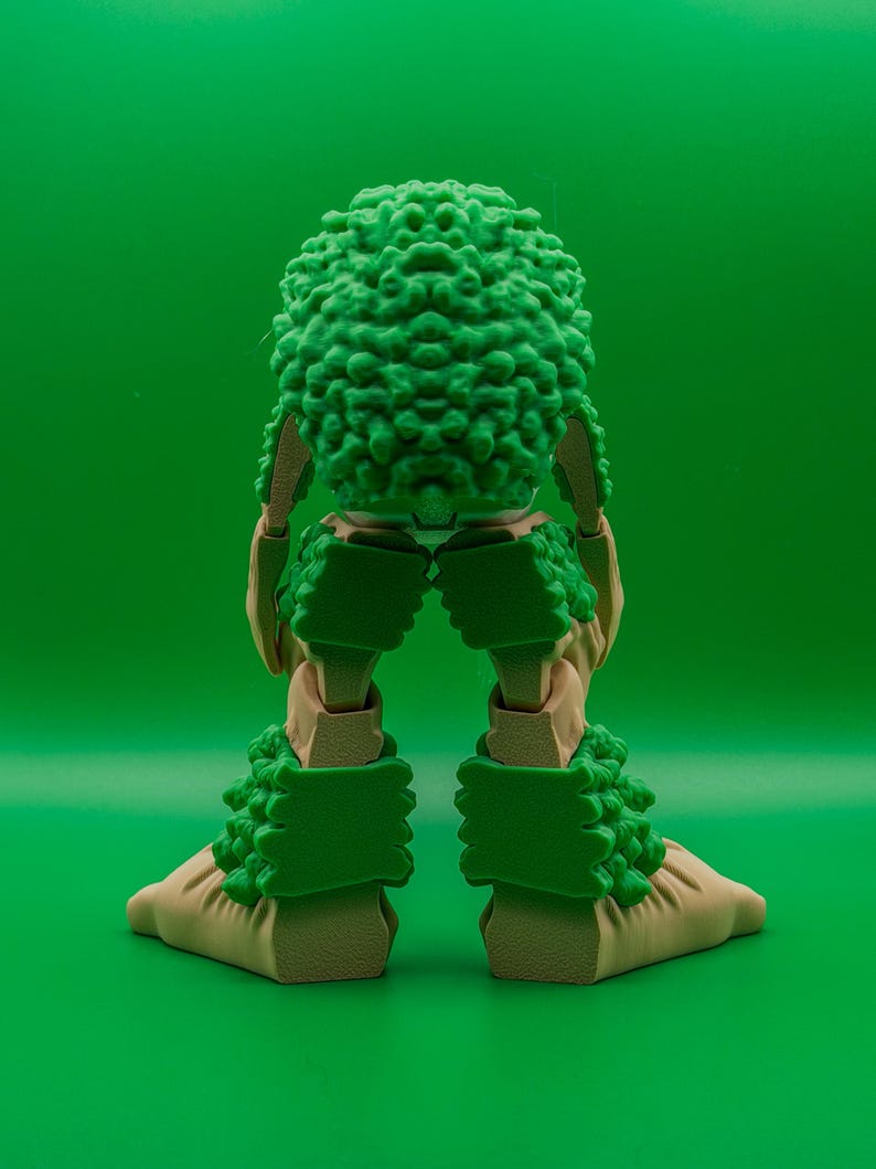 May include: An articulated figure in green and tan, featuring a textured, spherical head and matching leg and foot coverings. The limbs are tan, and the green elements have a textured, organic appearance. The background is a solid green.