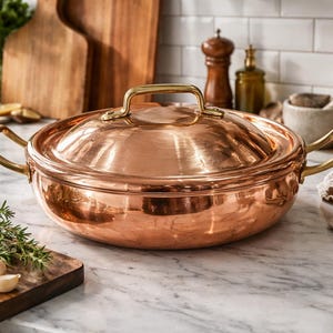 May include: A copper cooking pot with a lid and brass handles. The pot is round and reflective, sitting on a marble countertop. A wooden cutting board with herbs, garlic, and a lemon half is in the foreground. The pot is approximately 12 inches in diameter.
