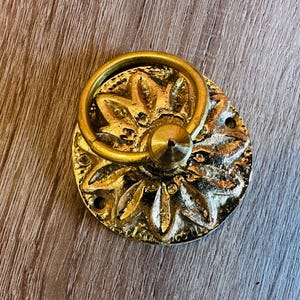 May include: A gold-toned door knocker with a circular design featuring a floral pattern and a ring handle.