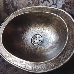 Op de afbeelding: Ovale, gehamerde metalen wastafel met ingewikkelde bloemen- en geometrische patronen. De wastafel heeft een donkere, verouderde afwerking en een decoratieve rand. De afvoer is gecentreerd, wat een rustieke uitstraling geeft.