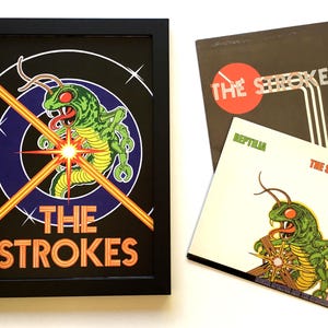 May include: A framed poster with a black background and a colourful graphic of a green and yellow bug with red eyes. The bug is surrounded by a blue and yellow circle with a starburst in the centre. The text "THE STROKES" is printed in orange below the bug.