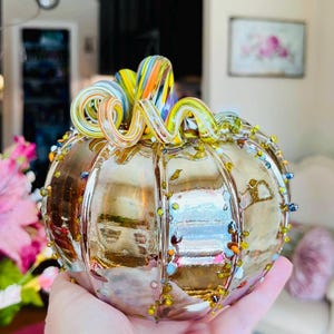 May include: A decorative glass pumpkin with a metallic gold finish and vertical ribbed segments. The top features a multi-colored, spiraled glass stem in shades of orange, yellow, and blue. Small, colorful glass beads are applied along the ribs.