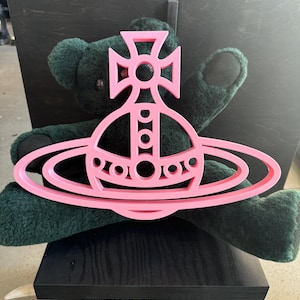 May include: A pink, flat, orb-shaped emblem featuring a cross at the top and a surrounding ring, positioned in front of a dark green, fuzzy teddy bear. The emblem has a cutout design with circular details along the central vertical axis.