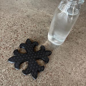 May include: A clear glass bottle filled with a clear liquid, next to a black, floral-shaped coaster. The coaster has a textured pattern. The items are on a textured, gray surface.