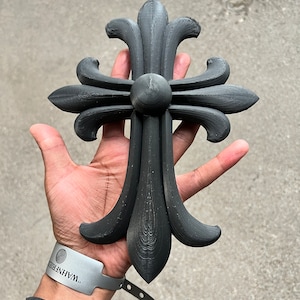 May include: A black, three-dimensional, ornate cross with a fleur-de-lis design. The cross is held in a person's hand.