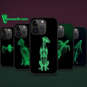 Puede incluir: Fundas de teléfono negras con diseños de rayos X verde brillante de un murciélago, gato, perro, tortuga y ballena. Las fundas tienen un acabado brillante y un recorte para la cámara. La marca "ImpressStudio" es verde.