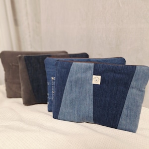 Könnte beinhalten: Eine Reihe von vier rechteckigen Reißverschlusstaschen, die in einer Linie angeordnet sind. Die vordere Tasche zeigt ein Patchwork-Design aus hell- und dunkelblauem Jeansstoff mit einem kleinen weißen rechteckigen Etikett, das ein Schmetterlingssymbol und den Text Alma Verde zeigt.