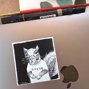 May include: A black and white illustration of a squirrel wearing a t-shirt with the word "RESIST" printed on it. The artwork is on a square, with the artist's signature in the lower left corner. A red paintbrush and a black marker are in the background.