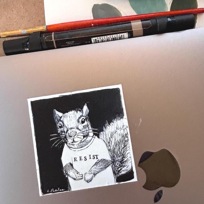 May include: A black and white illustration of a squirrel wearing a t-shirt with the word "RESIST" printed on it. The artwork is on a square, with the artist's signature in the lower left corner. A red paintbrush and a black marker are in the background.