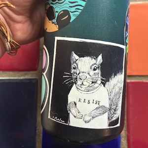 May include: A dark teal water bottle with several stickers. One sticker features a squirrel wearing a shirt with the word "RESIST" printed on it. The squirrel is in black and white, set against a black background.