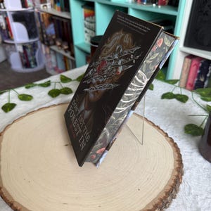 Peut inclure: Un livre intitulé "Don't Let the Forest In" est présenté sur une tranche de bois. La couverture du livre présente un fond sombre avec un motif de rose et d'épines. Le livre est posé sur un support transparent, mettant en valeur le dos et l'illustration de la couverture.