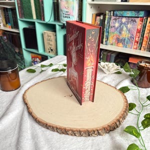 May include: A hardcover book titled "Daughter of the Worlds" with a red cover and gold lettering. The book is standing upright on a wooden slice, with a white cloth background and a bookshelf in the background.