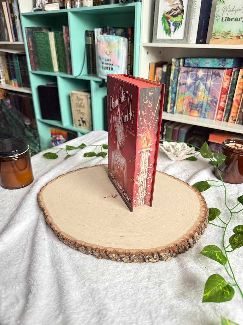 May include: A hardcover book titled "Daughter of the Worlds" with a red cover and gold lettering. The book is standing upright on a wooden slice, with a white cloth background and a bookshelf in the background.
