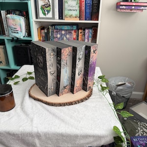 May include: Four hardback books with ornate covers are arranged on a wooden slice on a white cloth-covered table. The books feature silver and black designs, with visible titles and author names. A bookshelf with books and decorative items is in the background.