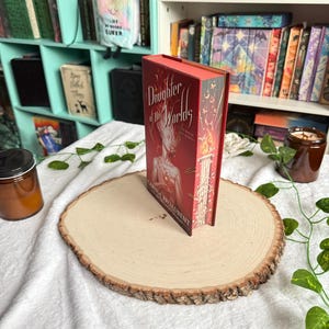 May include: A hardcover book titled "Daughter of the Worlds" with a red cover and gold lettering, standing upright on a wooden slab. The book's spine is visible, and the background includes a bookshelf and decorative elements.