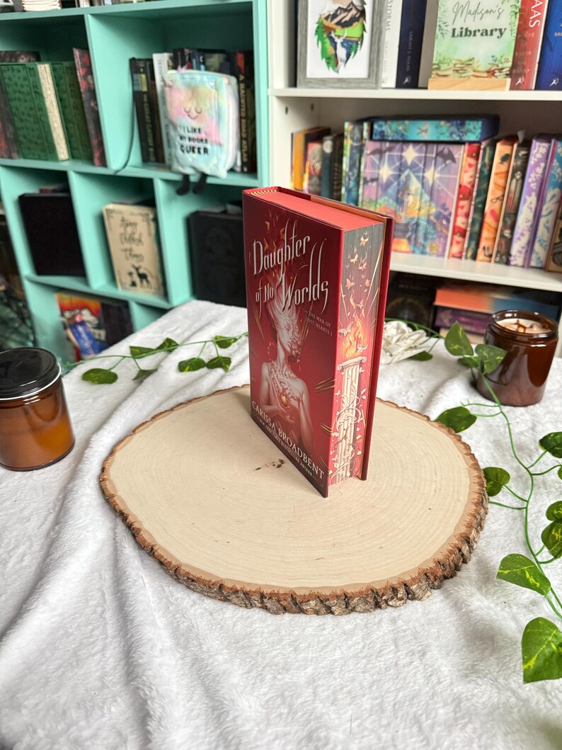 May include: A hardcover book titled "Daughter of the Worlds" with a red cover and gold lettering, standing upright on a wooden slab. The book's spine is visible, and the background includes a bookshelf and decorative elements.