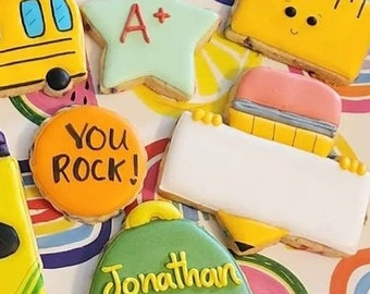 Op de afbeelding: Een set van negen gedecoreerde suikerkoekjes met een terug naar school thema. De koekjes bevatten een schoolbus, een appel, kleurpotloden, een liniaal, een rugzak, een schoolgebouw, een ster en twee gepersonaliseerde rugzakken. De koekjes zijn versierd met kleurrijk glazuur en hebben verschillende teksten, waaronder "You Rock!" en "Back to School".