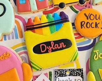 Op de afbeelding: Een verzameling gedecoreerde suikerkoekjes met een terug naar school thema. De koekjes bevatten een schoolbus, een ster, een liniaal, een potlood, een doos met kleurpotloden, een appel en twee rugzakken. De koekjes zijn versierd met kleurrijk glazuur en hebben gepersonaliseerde namen. De tekst "Happy First Day" en "Back to School" zijn ook inbegrepen.