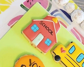 Op de afbeelding: Een verzameling versierde koekjes met een schoolthema. De koekjes omvatten een rood schoolgebouw, een gele schoolbus, een liniaal, een ster met een "A+" en een rond koekje met "YOU ROCK!" erop. De koekjes liggen op een kleurrijke achtergrond.