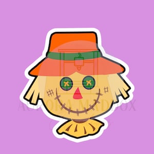 May include: A cartoon scarecrow sticker with a smiling face, button eyes, and a stitched mouth. The scarecrow wears an orange hat with a green band and has straw-like hair. The background is a solid lavender colour.