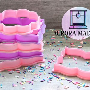 May include: A set of four pink and purple plastic cookie cutters shaped like a scalloped rectangle. The cutters are stacked on top of each other. The cutters are on a wooden surface with colorful sprinkles.