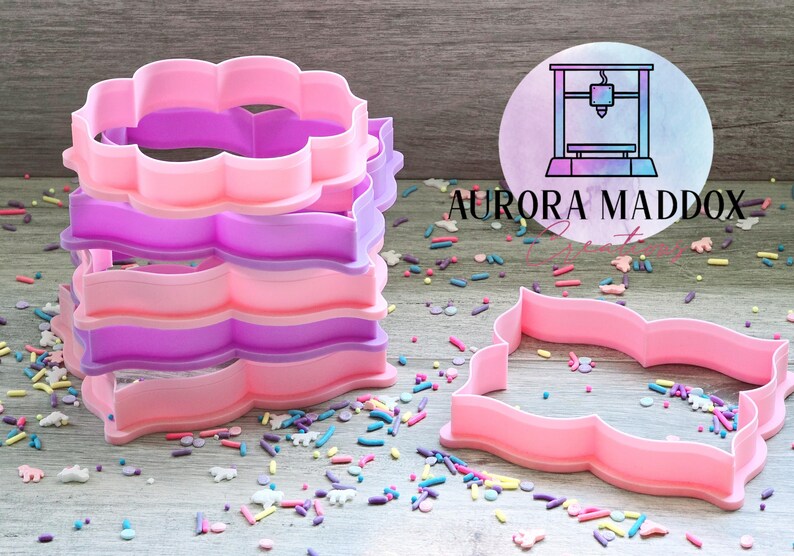 May include: A set of four pink and purple plastic cookie cutters shaped like a scalloped rectangle. The cutters are stacked on top of each other. The cutters are on a wooden surface with colorful sprinkles.