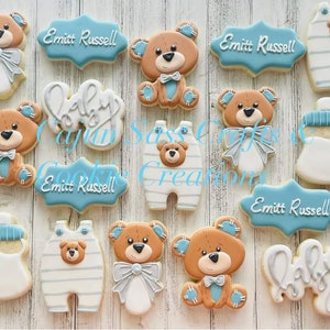K&ouml;nnte beinhalten: Ein Set mit 12 dekorierten Zuckerkeksen mit einem Babyb&auml;ren-Motiv. Die Kekse sind mit blauem, wei&szlig;em und braunem Zuckerguss verziert. Die Kekse beinhalten eine Babyflasche, einen Baby-Strampler und einen Teddyb&auml;r mit Fliege. Die Kekse sind mit dem Text "Baby" und "Emmitt Russell" verziert.