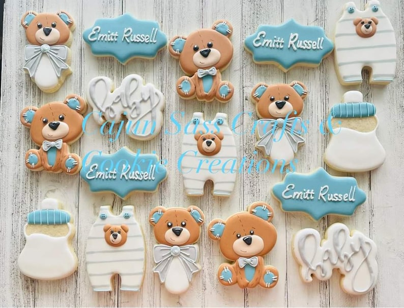 K&ouml;nnte beinhalten: Ein Set mit 12 dekorierten Zuckerkeksen mit einem Babyb&auml;ren-Motiv. Die Kekse sind mit blauem, wei&szlig;em und braunem Zuckerguss verziert. Die Kekse beinhalten eine Babyflasche, einen Baby-Strampler und einen Teddyb&auml;r mit Fliege. Die Kekse sind mit dem Text "Baby" und "Emmitt Russell" verziert.