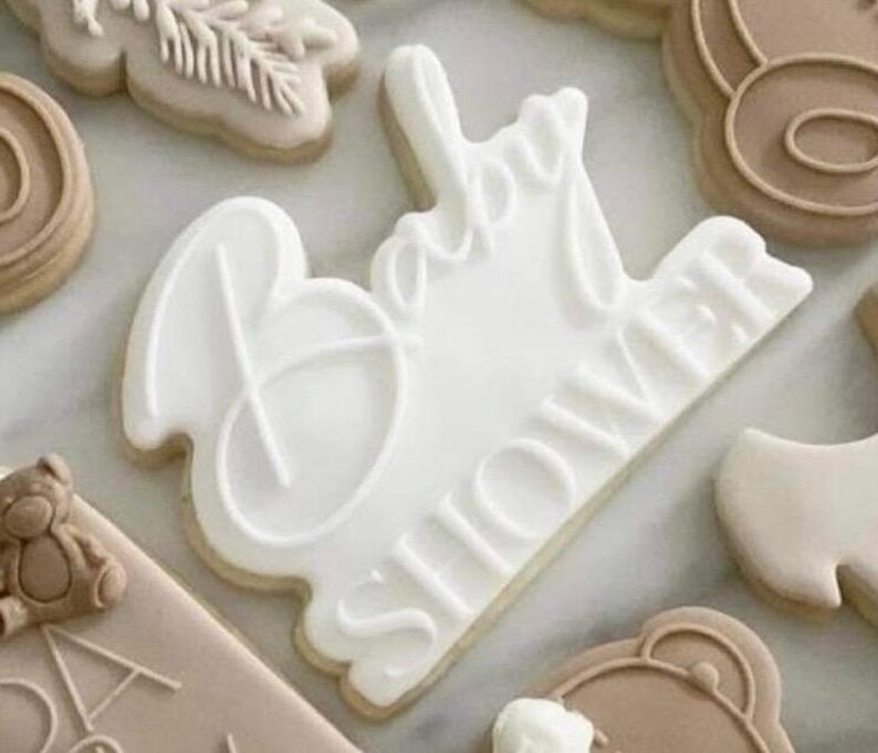 May include: White cookie with the words "Baby Shower" in cursive script. The cookie is decorated with white icing.