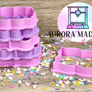 May include: A set of four pink and purple plastic cookie cutters in the shape of a scalloped rectangle. The cutters are stacked on top of each other and are surrounded by colorful sprinkles.