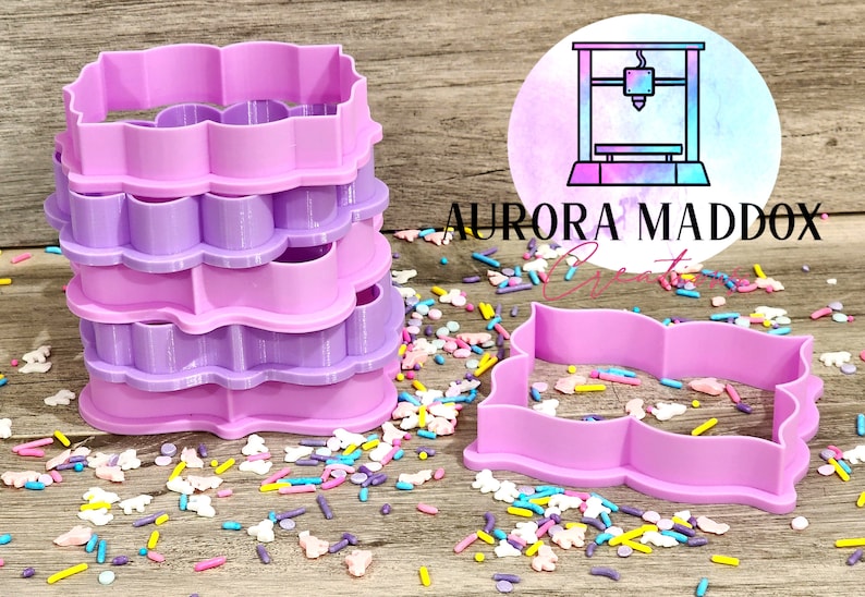 May include: A set of four pink and purple plastic cookie cutters in the shape of a scalloped rectangle. The cutters are stacked on top of each other and are surrounded by colorful sprinkles.