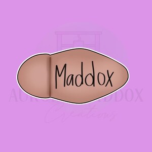 Puede incluir: Un cortador de galletas marrón en forma ovalada con el nombre "Maddox" en escritura negra. El cortador de galletas tiene un contorno blanco y está sobre un fondo morado claro.