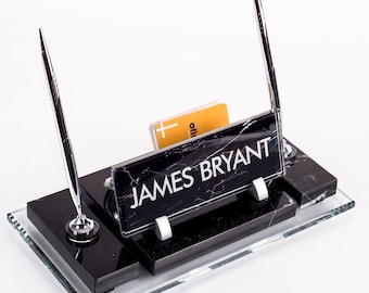 Könnte beinhalten: Schwarzer Marmor-Schreibtisch-Organizer mit Glasbasis. Der Organizer verfügt über einen silbernen Stifthalter, einen Visitenkartenhalter und ein Namensschild mit der Aufschrift "JAMES BRYANT".